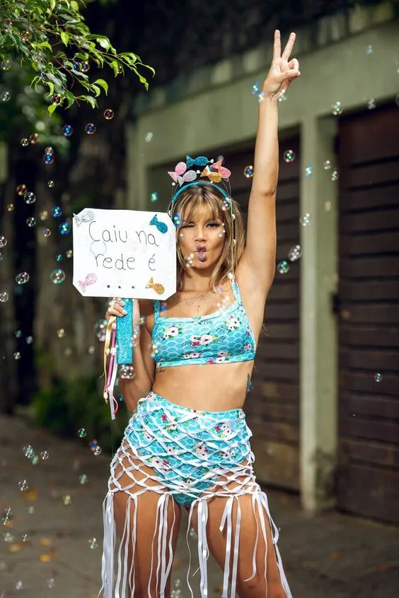 Mulher de pele clara com top e hot pant azuis, tiara de peixes e placa escrito "caiu na rede é..."