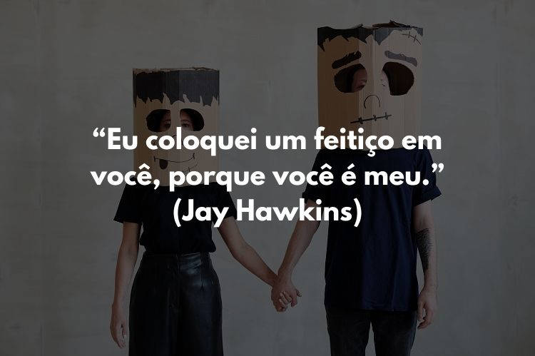 Foto de duas pessoas de mãos dadas, usando roupas pretas e caixa de papelão na cabeça. Ao centro, frase "Eu coloquei um feitiço em você, porque você é meu.” (Jay Hawkins)
