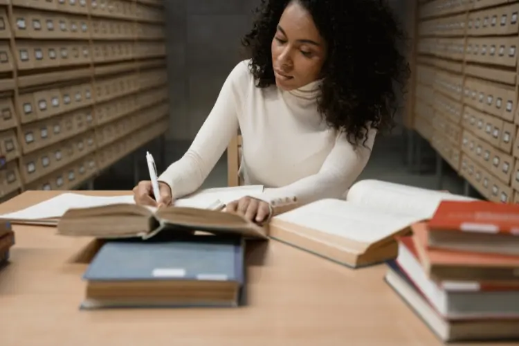 Mulher de pele negra escrevendo em caderno em meio aos livros e arquivos