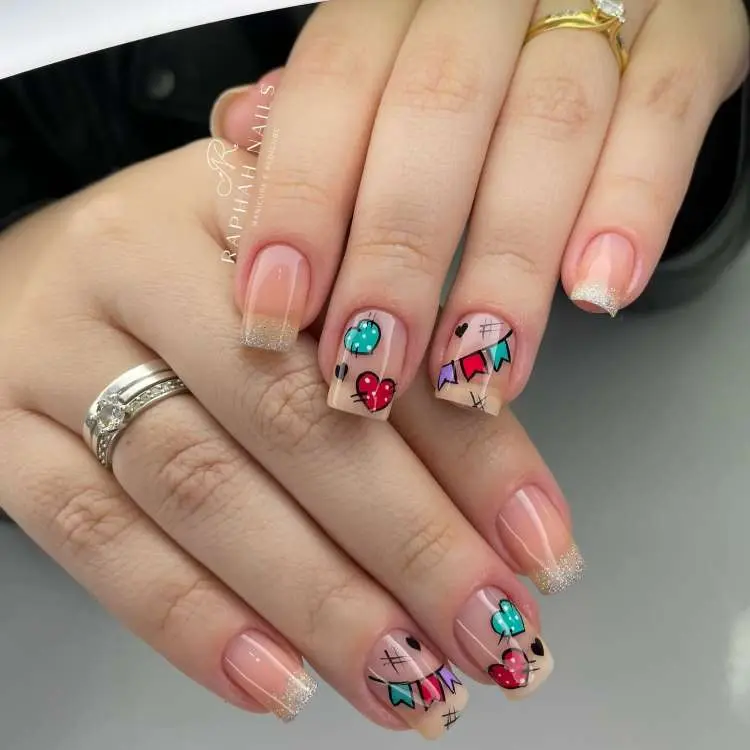 manicure com unhas de formato quadrado e pontas com glitter prateado. Algumas unhas exibem desenhos vibrantes de corações, balões e bandeirinhas coloridas, reminiscentes de festas juninas. A mão ostenta anéis, incluindo um com uma pedra verde.