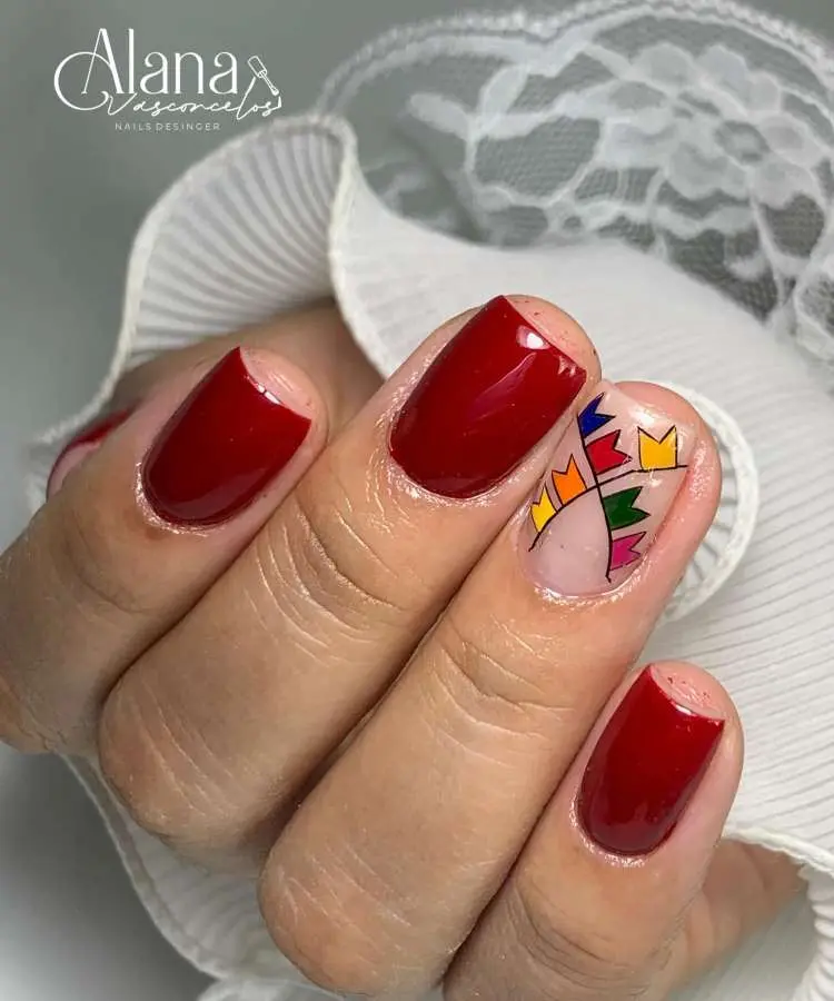 Mão com unhas curtas e escuras em vermelho vinho. Uma unha tem um desenho de bandeirinhas de Festa Junina em tons de amarelo, azul e vermelho sobre base clara.