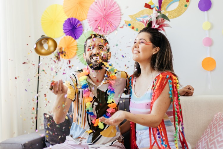 Homem e mulher de pele clara com roupas carnavalescas fazendo festa em casa, com decorações da folia, glitter, confete e serpentina