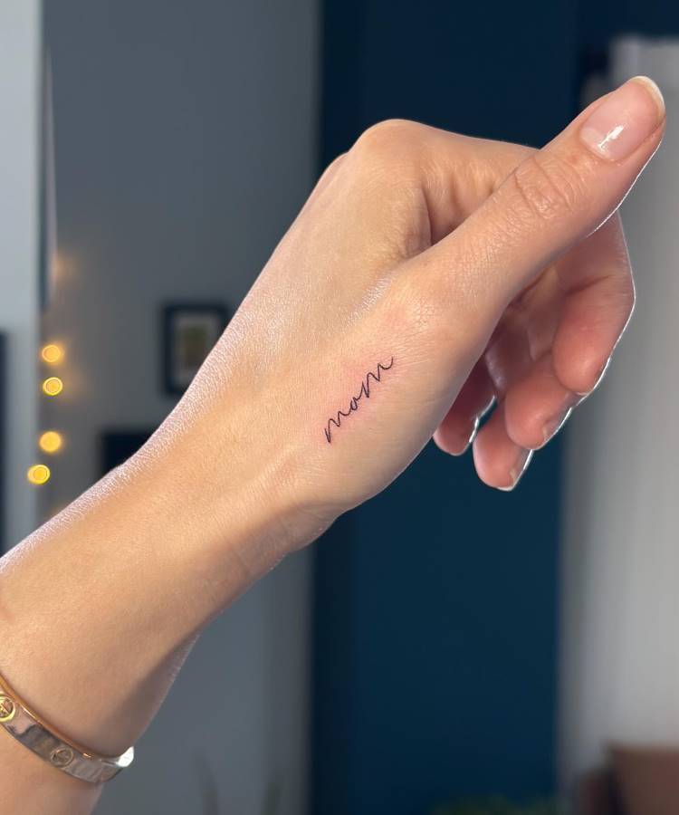 A imagem mostra a parte superior da mão de uma pessoa, com uma pequena tatuagem minimalista e fina da palavra "mom" escrita em cursiva preta, posicionada ao longo da linha do dedo mindinho. A pessoa usa uma pulseira dourada. O fundo está desfocado.








