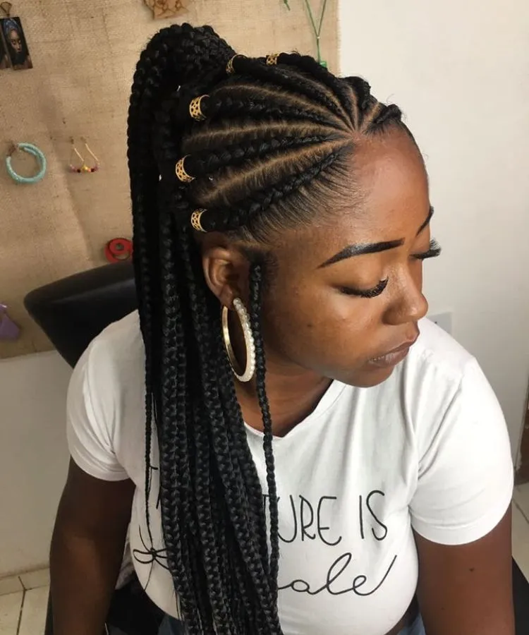 Mulher negra com trança nagô, penteado rabo de cavalo, com joias. Ela usa camiseta branca com frase e brinco de argola com pérolas