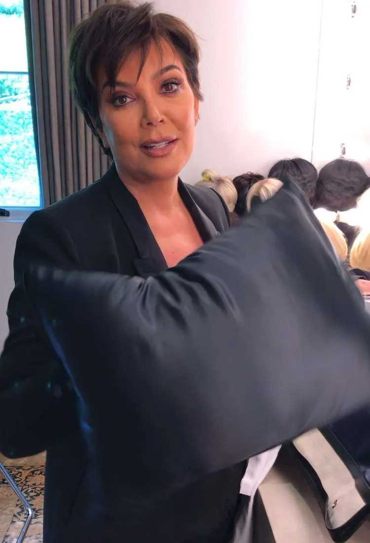Kris Jenner sorri para a câmera enquanto segura uma almofada escura. Ela veste um blazer preto e seu cabelo curto e escuro é levemente espetado, em um ambiente interno com perucas ao fundo.