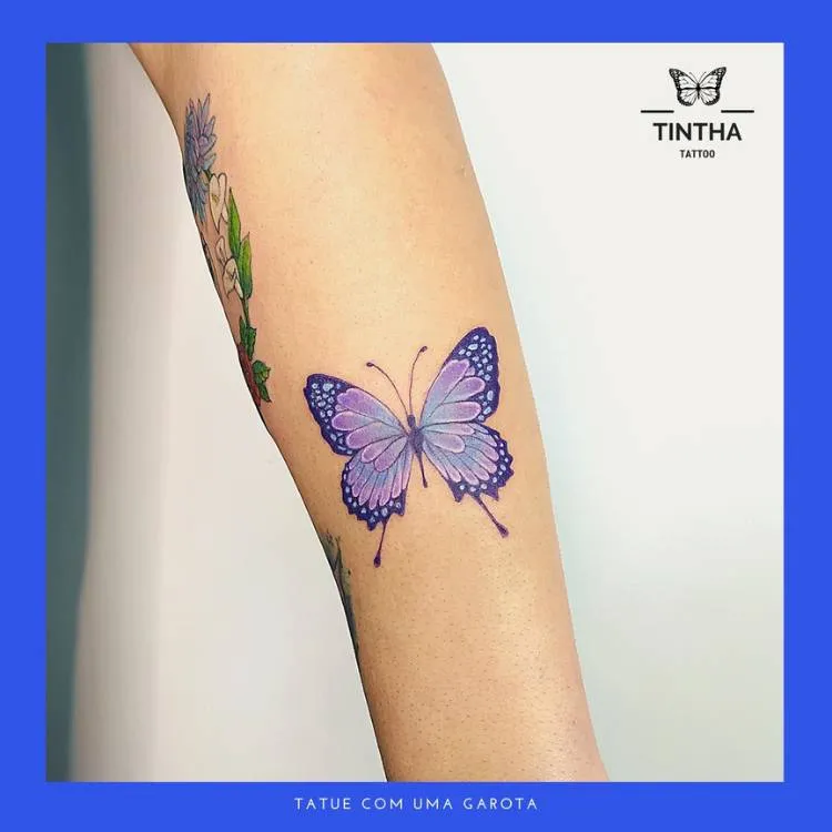 Tatuagem de borboleta em traços finos com tons de azul e roxo no braço de uma mulher de pele clara. 