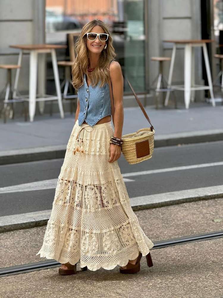 Mulher de pele clara de colete jeans e saia longa de crochê, com bolsa de palha e salto marrom