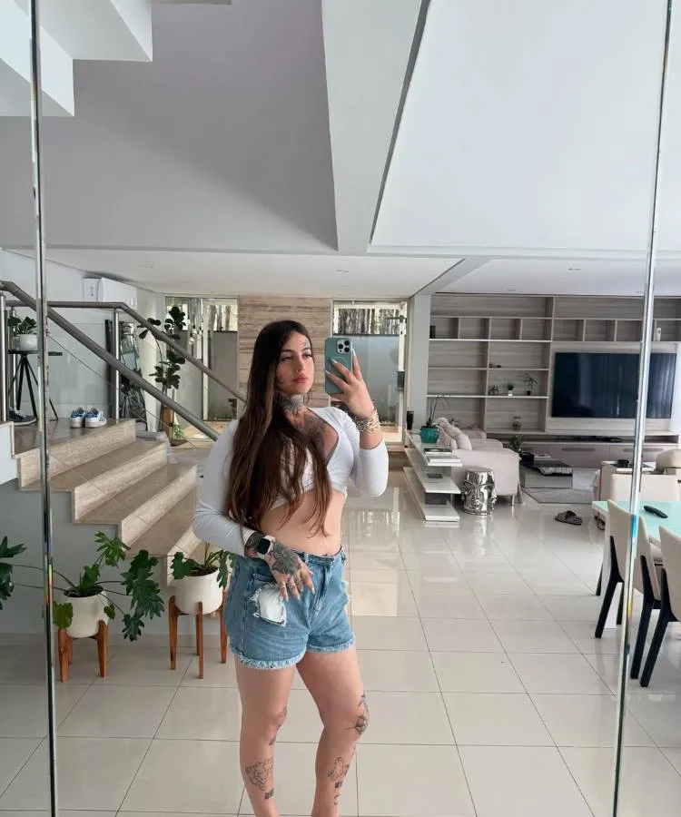 Mulher fotografa corpo inteiro em espelho de sala ampla e moderna. Usa cropped branco e shorts jeans, revelando tatuagens nas pernas e braços. Cabelos longos soltos, expressão séria. Ambiente luxuoso ao fundo exibe escada, plantas, mesa de jantar e sofá.
