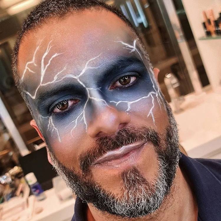 Homem de pele negra com maquiagem azul cintilante na região dos olhos, com raios brancos 