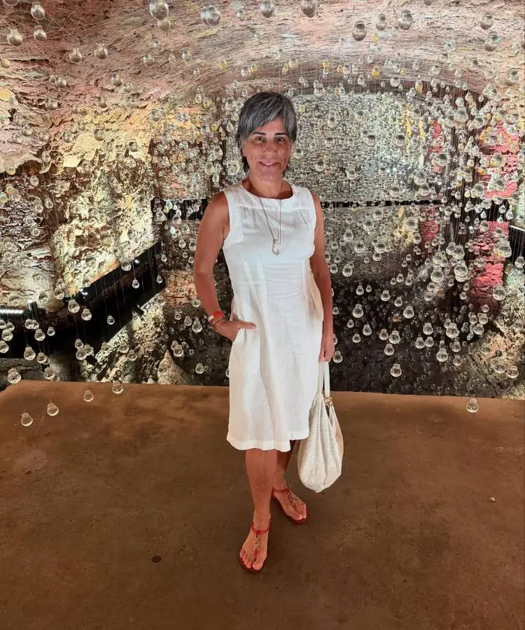 Mulher de cabelos curtos e grisalhos veste um vestido branco e sandálias vermelhas. Segura uma bolsa clara ao lado. Fundo com instalação artística composta por pequenas esferas suspensas e parede texturizada, criando um ambiente encantador e único.