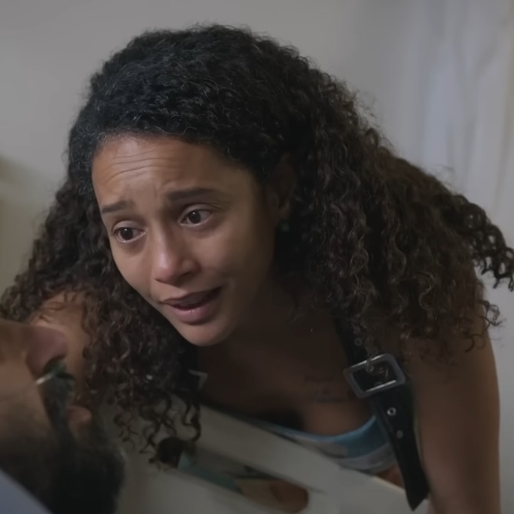 Taís Araújo como Raquel em Vale Tudo. Mulher com cabelo cacheado e expressão emocionada conversa inclinada sobre uma pessoa deitada em uma cama de hospital. Ela veste alças pretas de macacão, em ambiente claro com fundo desfocado, transmitindo intensidade e preocupação.





