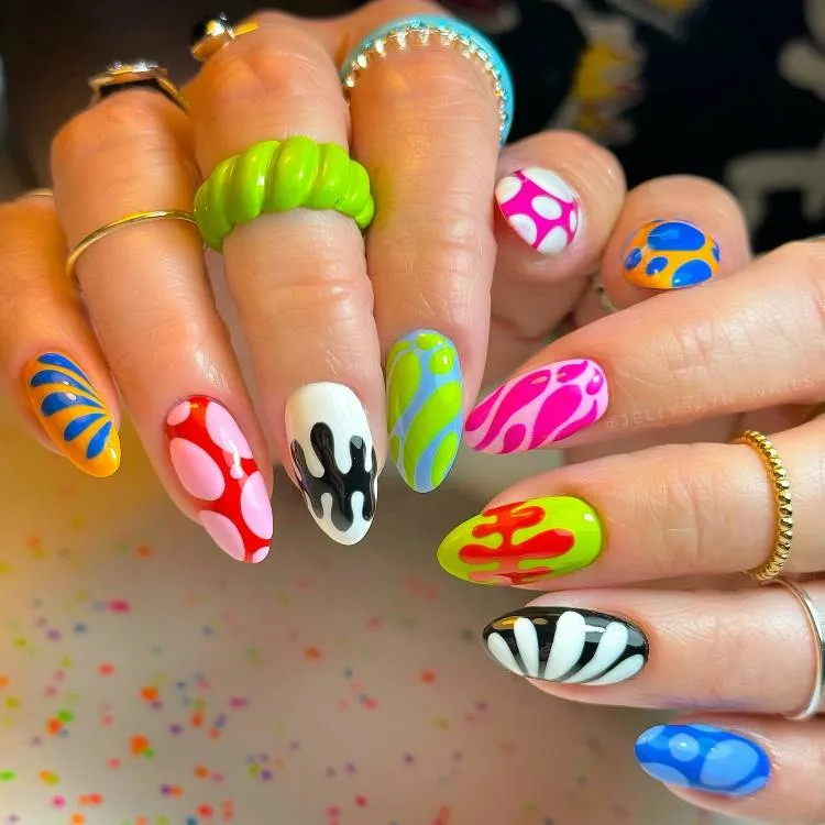 A imagem mostra um close-up de duas mãos com os dedos estendidos, exibindo uma variedade de designs de arte nas unhas. Cada unha tem um padrão e esquema de cores diferentes. Os designs incluem formas abstratas, redemoinhos, pontos e listras em cores vibrantes como azul, verde, amarelo, rosa, vermelho, preto e branco. O fundo está desfocado, com manchas coloridas que sugerem um ambiente festivo ou relacionado a cuidados de beleza.