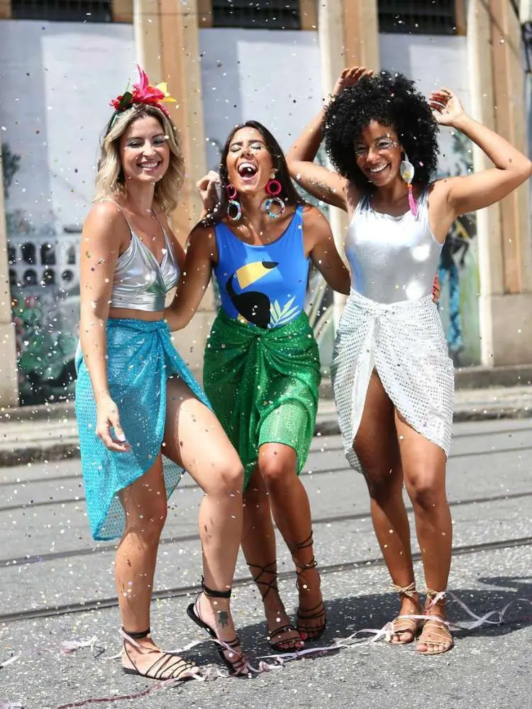 Três mulheres usando saia de lantejoula, sendo uma com top prata, outra com body azul de tucano e outra de body prata 