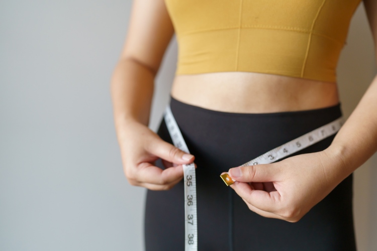 Mulher de pele clara usando top amarelo e calça preta medindo cintura com fita métrica
