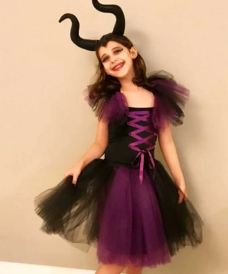Garota com fantasia de bruxa infantil Malévola. Cores predominantes: . Usa: vestido + tiara de chifres da personagem