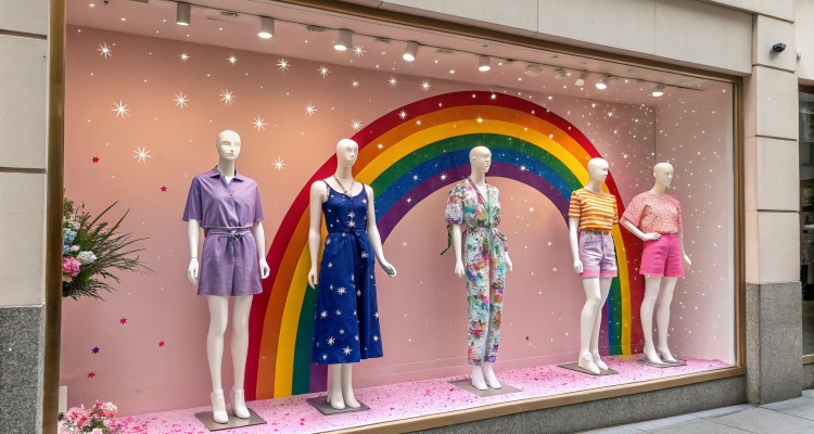 Vitrine com cinco manequins usando roupas coloridas, dispostas em frente a uma parede rosa decorada com um grande arco-íris e estrelas brilhantes. Chão coberto de glitter rosa, flores à esquerda, iluminação suave cria um ambiente vibrante e alegre.
