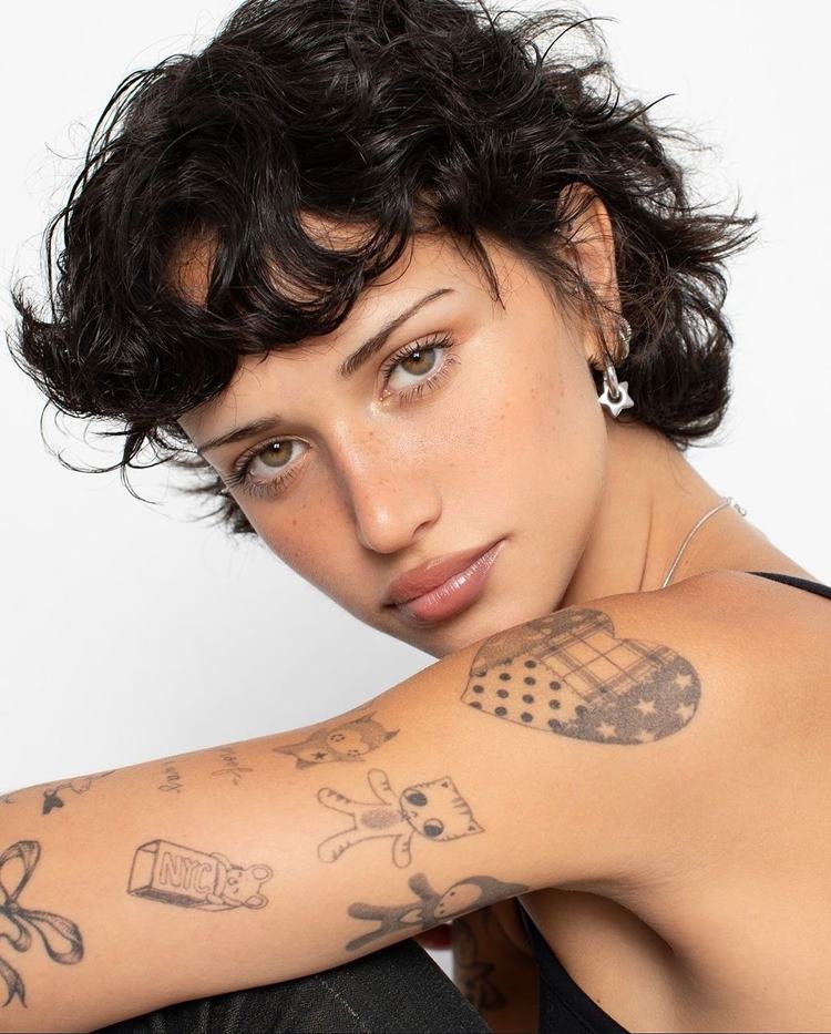 Mulher de cabelos cacheados curtos, com tatuagens no braço, incluindo desenhos de gatos e um ursinho. Ela está vestindo regata preta e brincos delicados, com expressão suave e olhar direto. O fundo é branco, destacando seu estilo moderno e artístico.