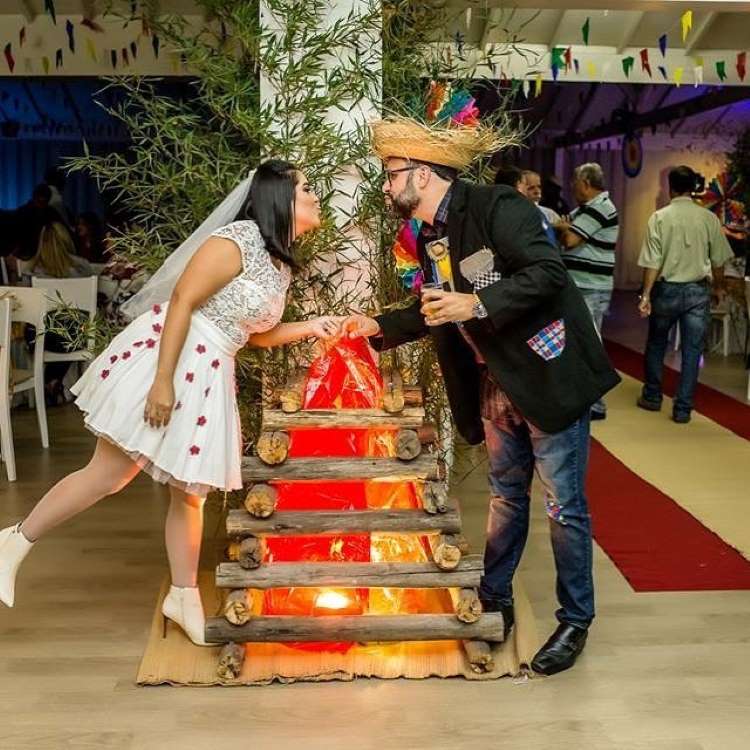 Um casal com trajes festivos compartilha um momento divertido ao lado de uma fogueira decorativa. A mulher, em um vestido branco com detalhes de flores vermelhas e véu, se inclina para beijar o homem, que usa chapéu de palha e jaqueta xadrez. O ambiente interno é decorado com bandeiras coloridas, sugerindo uma celebração animada.
Fontes