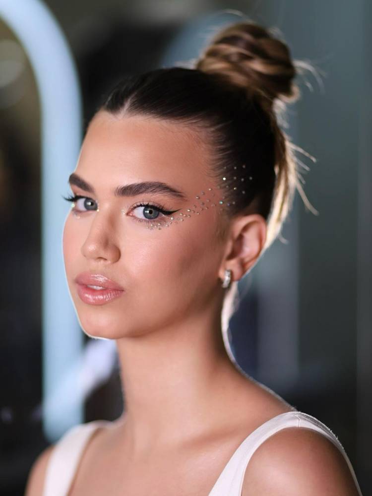 Mulher de pele clara com strass no rosto e coque alto. 