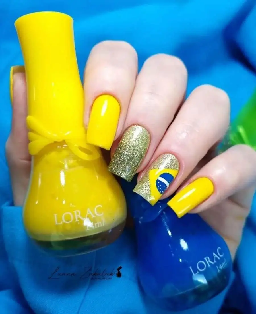 Imagem de mão com unhas decoradas de Brasil.  Cores usadas, amarelo, azul, branco e glitter dourado. Polegar: amarelo. Indicador: amarelo. Dedo Médio, glitter dourado. Anelar: glitter dourado + bandeira do Brasil. Mínimo: amarelo.