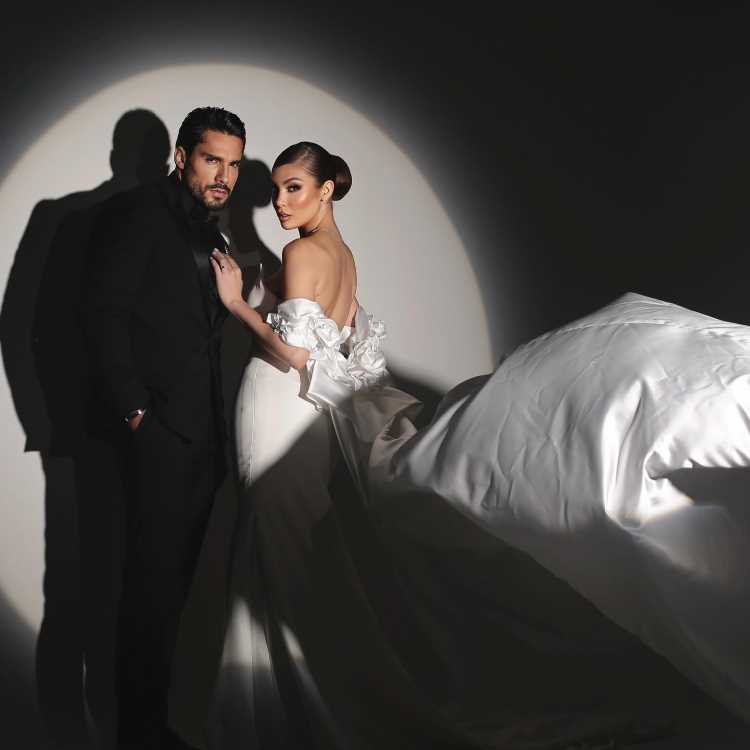Casal Bil Araújo e Adriana Yanca posa sob luz dramática, em ambiente sofisticado. A noiva veste um vestido branco com detalhes florais e cauda fluida, enquanto o noivo usa terno preto elegante. A composição realça modernidade, glamour e romantismo da fotografia.
