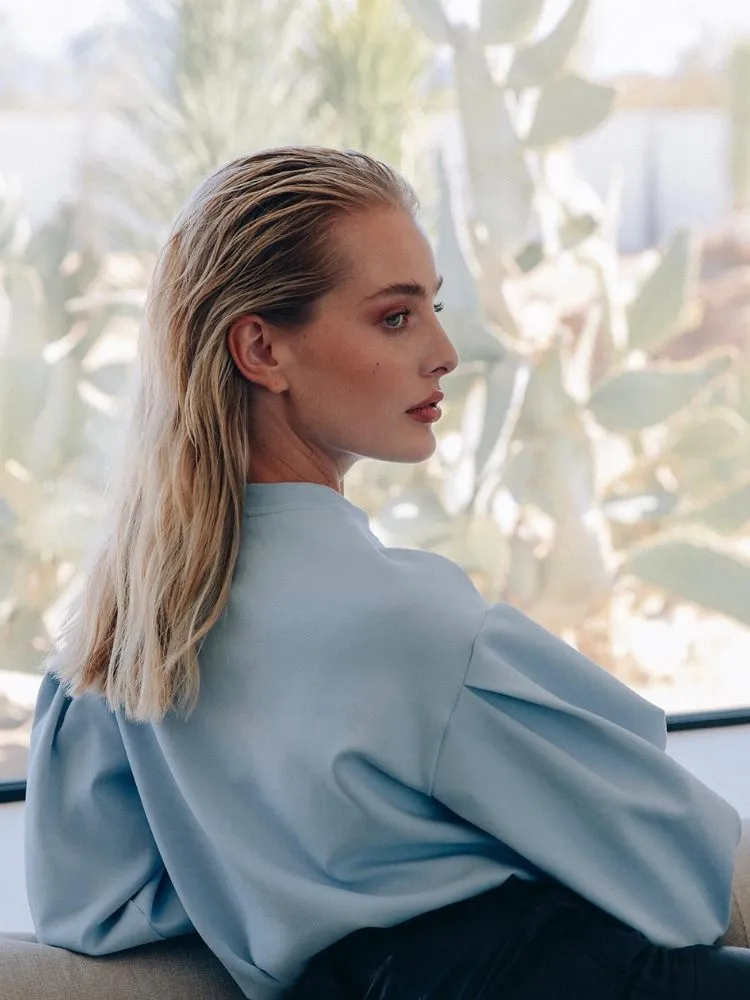 Mulher de pele clara em frente à janela com cabelo loiro estilo slick back e camisa azul