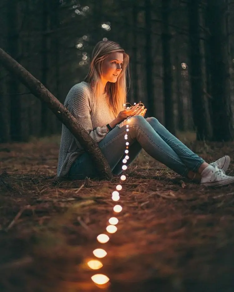 Frase para foto pensativa mulher branca loira no meio da floresta a noite, sentada segurando luzes veste calça jeans, tênis e tricot bege Frase para foto pensativa