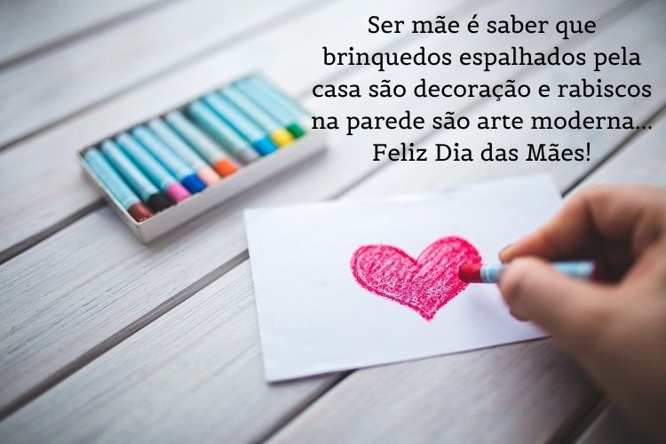 Foto de pessoa de pele clara desenhando coração com giz de cera rosa e frase " Ser mãe é saber que brinquedos espalhados pela casa são decoração e rabiscos na parede são arte moderna… Feliz Dia das Mães!"