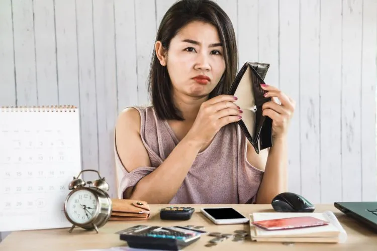 Uma mulher com uma expressão de descontentamento segura uma carteira vazia, sentada em uma mesa com um calendário, celular e calculadora. A imagem sugere uma preocupação financeira e a necessidade de estratégias para sair do vermelho.

