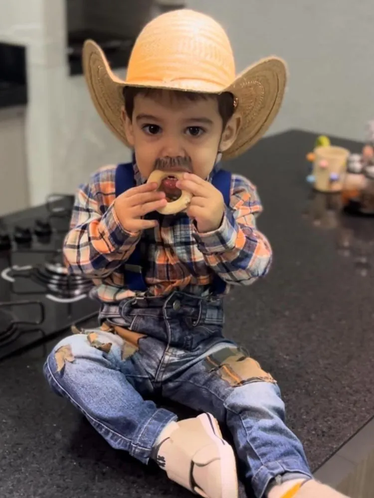 Menino de pele clara comendo biscoito usando chapéu de palha country, camisa xadrez, suspensório e calça jeans com retalho 