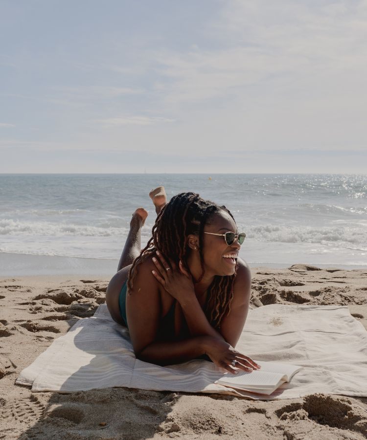 Uma mulher negra fazendo bronzeado saudável na praia, sob uma canga bege. Ela usa biquíni verde, óculos de sol, está de bruços e apoia uma das mãos em um livro na canga.