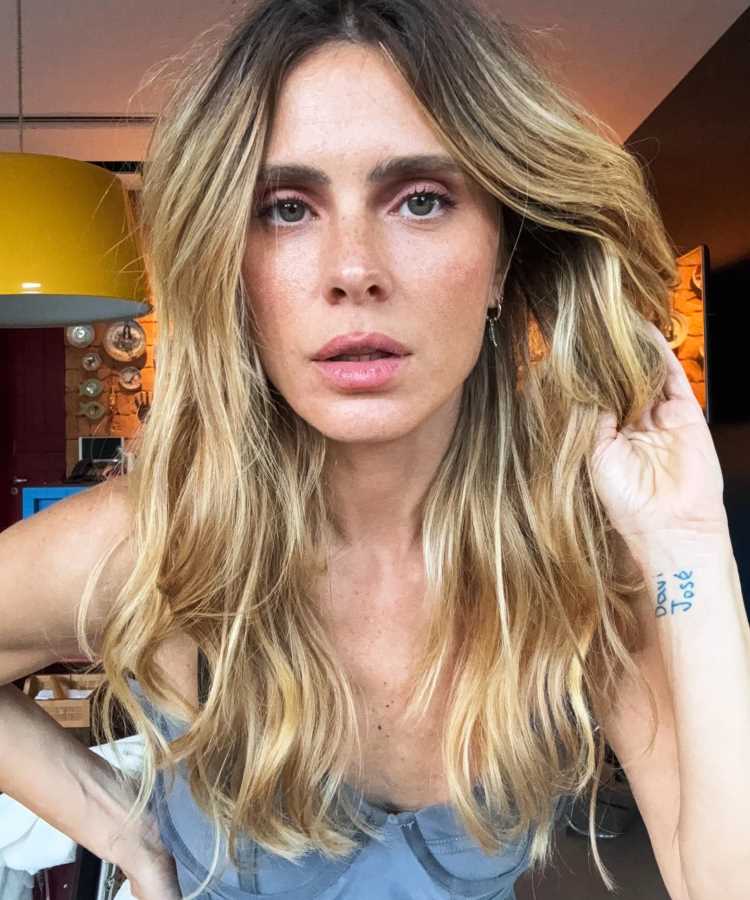 Carolina Dieckmann loira com cabelos longos e ondulados posa em ambiente interno com iluminação quente. Ela veste blusa cinza, exibe expressão natural e tattoo discreta no braço. Frequentemente mencionada entre famosos que não envelhecem, sua beleza chama atenção.
