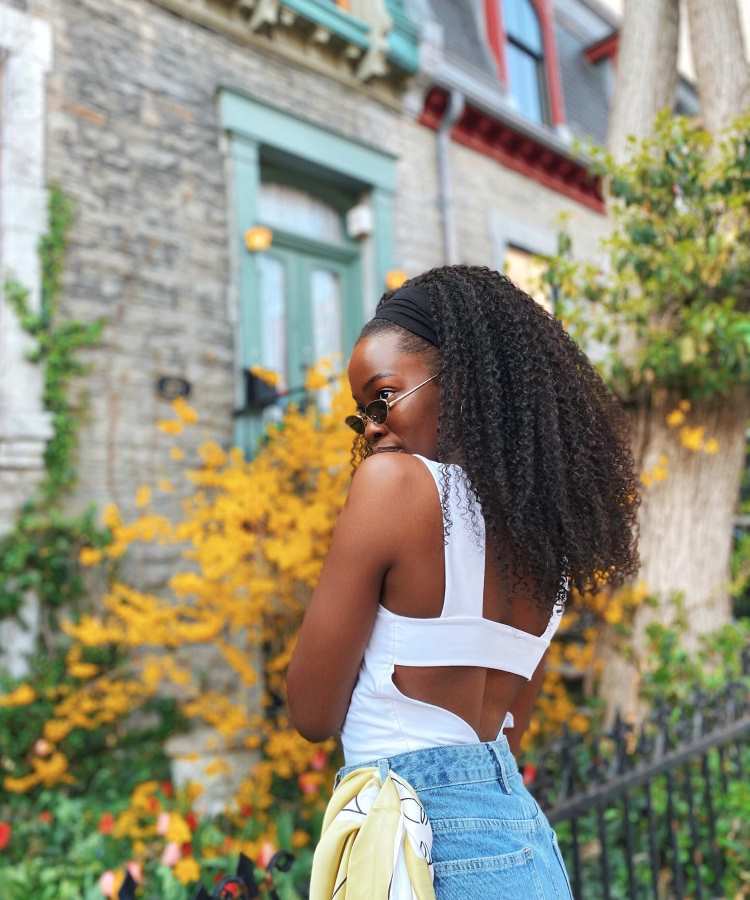 Mulher com cabelo cacheado volumoso solto, faixa preta, óculos escuros, blusa branca e jeans, posando ao ar livre com arbustos amarelos ao fundo.