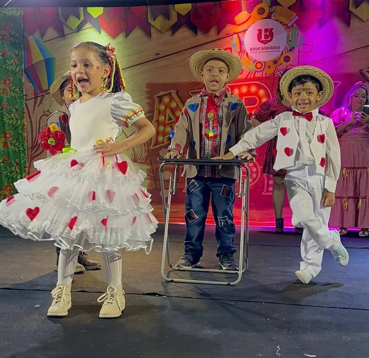 Uma menina e dois meninos fantasiados para quadrilha em um palco de arraial escolar.
