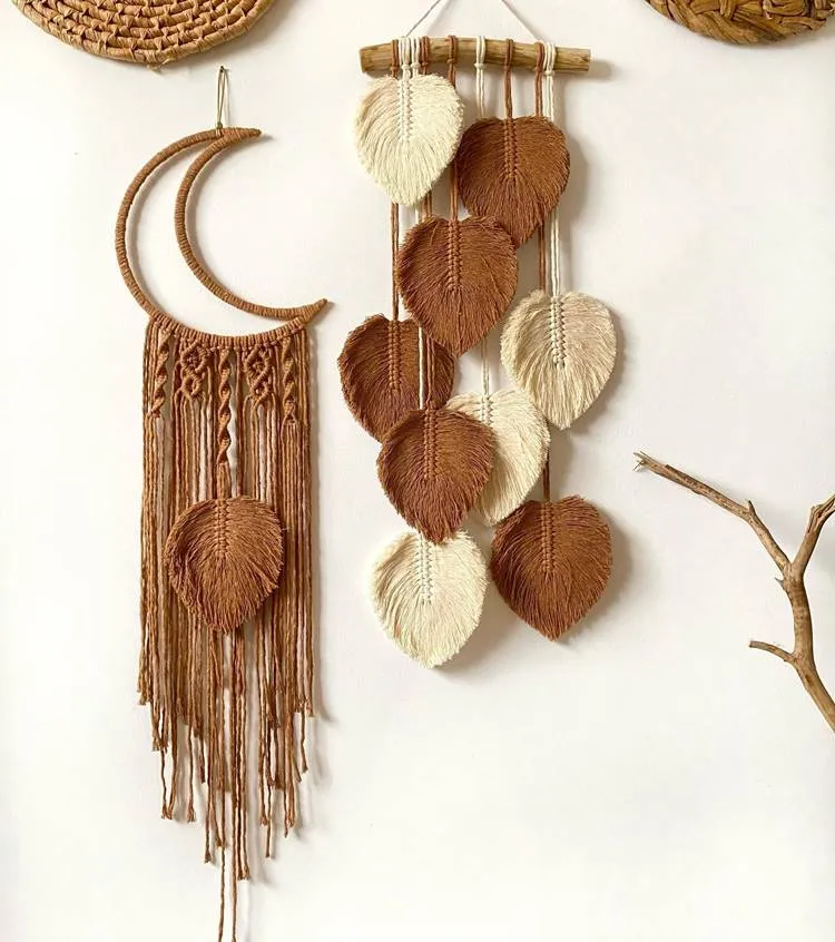 dois itens decorativos pendurados em uma parede branca. À esquerda, há um enfeite em forma de lua crescente feito de material entrelaçado marrom, com franjas penduradas na parte inferior. À direita, um enfeite retangular exibe folhas tecidas marrons e brancas penduradas verticalmente. Ambos os itens têm uma estética natural e boêmia. No canto superior direito da imagem, parte de outro item decorativo tecido pode ser visto. Um pequeno galho seco está posicionado diagonalmente no canto inferior direito da imagem.