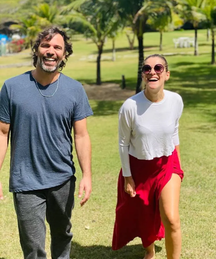Primos caminha e sorri em um gramado verde, com palmeiras ao fundo. O homem, com barba, usa uma camiseta azul e calça cinza. A mulher, de óculos de sol, usa uma blusa branca e saia vermelha. famosos que são parentes