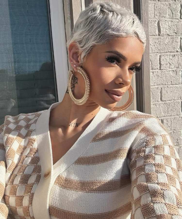 Mulher com cabelos curtos platinados e ondulados, usando brincos grandes dourados, maquiagem leve com cílios destacados, tatuagem no pescoço. Veste cardigan bege listrado e posa sob luz natural, com expressão serena e confiante ao ar livre.