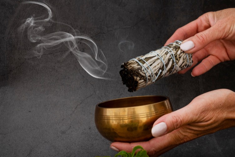 Foto de fundo escuro com pessoa de pele clara segurando pote dourado e incenso de sálvia queimando