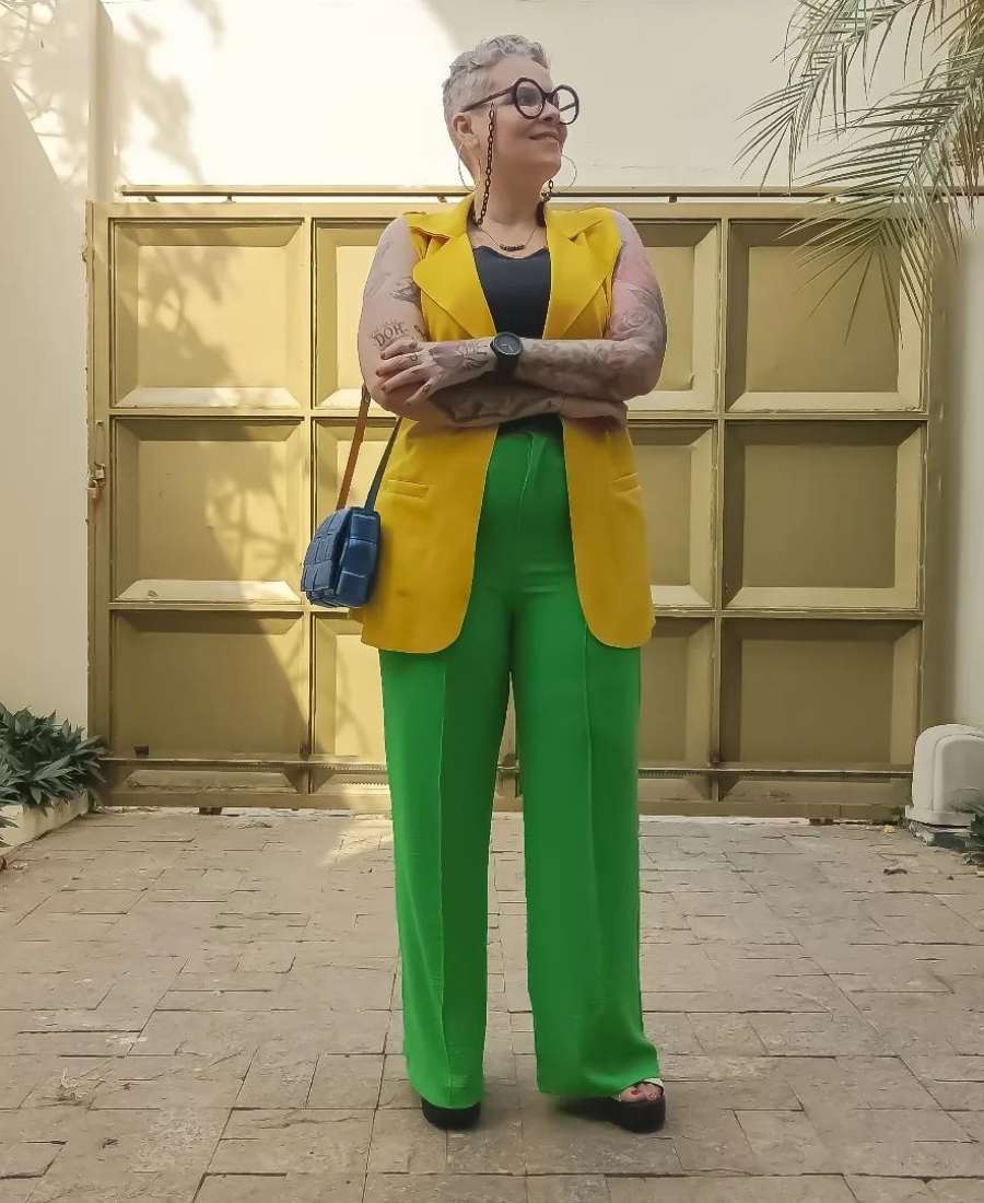 Imagem com fundo de portão de casa. No centro, mulher usando roupas Brazilcore: camiseta preta + calça verde + colete amarelo + bolsa azul.