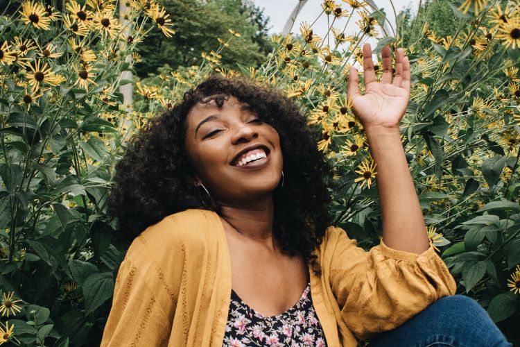 Mulher sorrindo alegremente em meio a flores amarelas, com roupa descontraída e expressão de felicidade. O cenário natural transmite leveza, harmonia e conexão com a natureza, com tons vibrantes e atmosfera calorosa em um dia ensolarado.