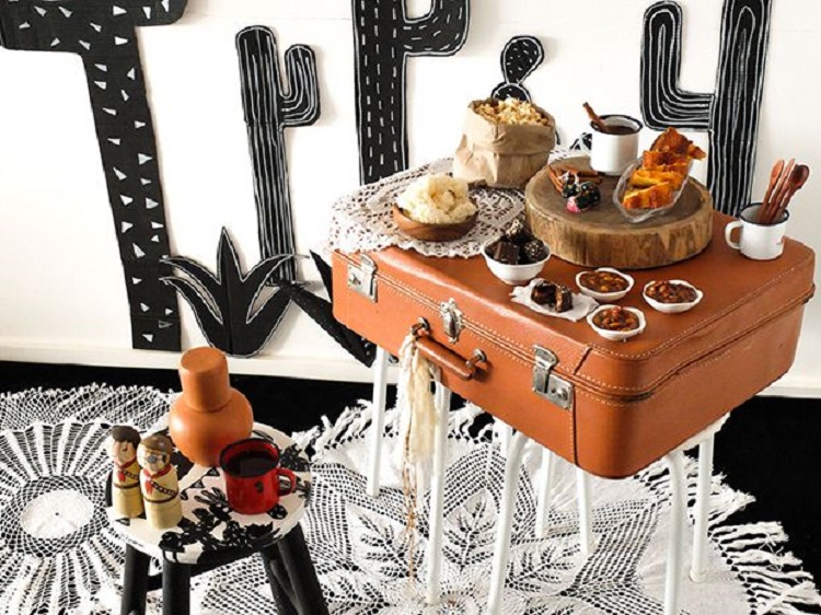 A imagem representa decoração para festa junina com mesas diferentes, em formato de mala. Contém doces, cactos, etc