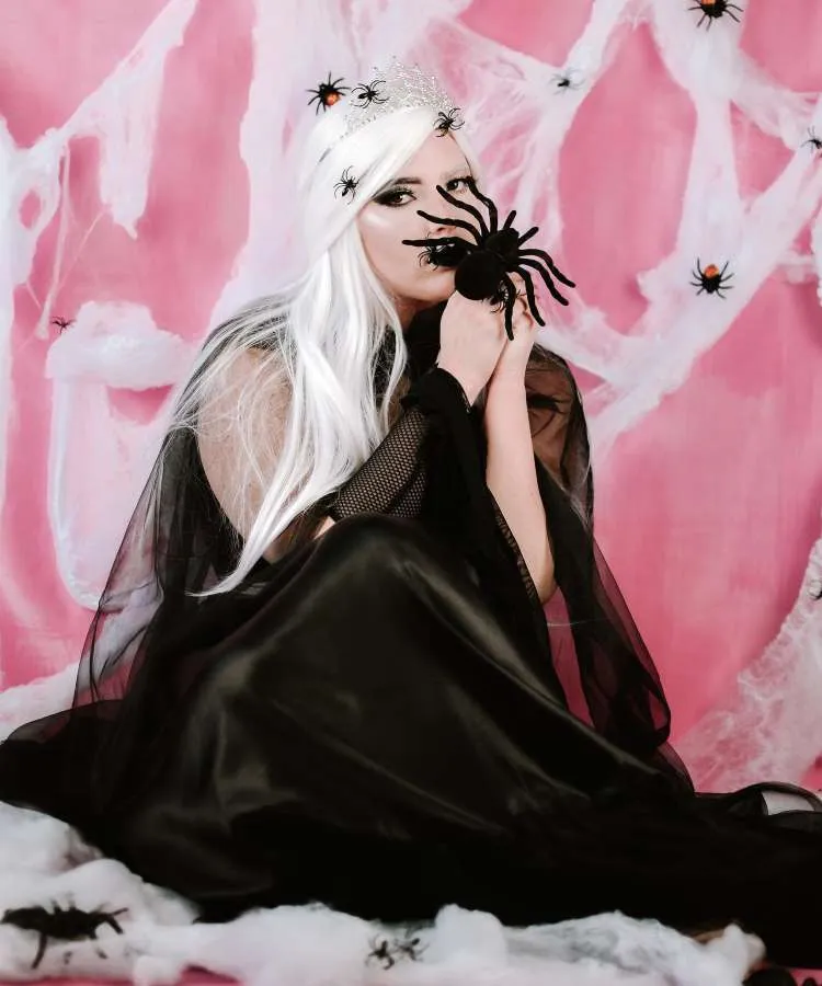 Mulher de pele clara usando peruca ranca, aranhas no cabelo, coroa e fantasia preta com capa de teia 