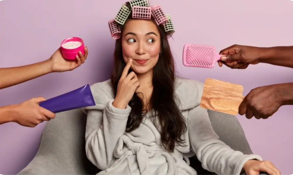 Mulher de pele clara usando roupão cinza, sentada em poltrona cinza, com bobs coloridos no cabelo e mãos oferecendo produtos como creme, escova de cabelo e espelho 