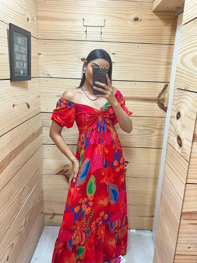 Mulher de pele clara tirando foto no espelho usando vestido longo estampado com fundo vermelho.