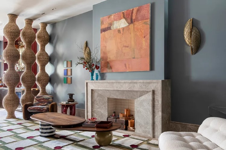A sala possui estilo estilizado com lareira de mármore, tapete geométrico, móveis contemporâneos e decoração étnica. Destacam-se colunas de vime, pintura abstrata e elementos decorativos nas paredes.

