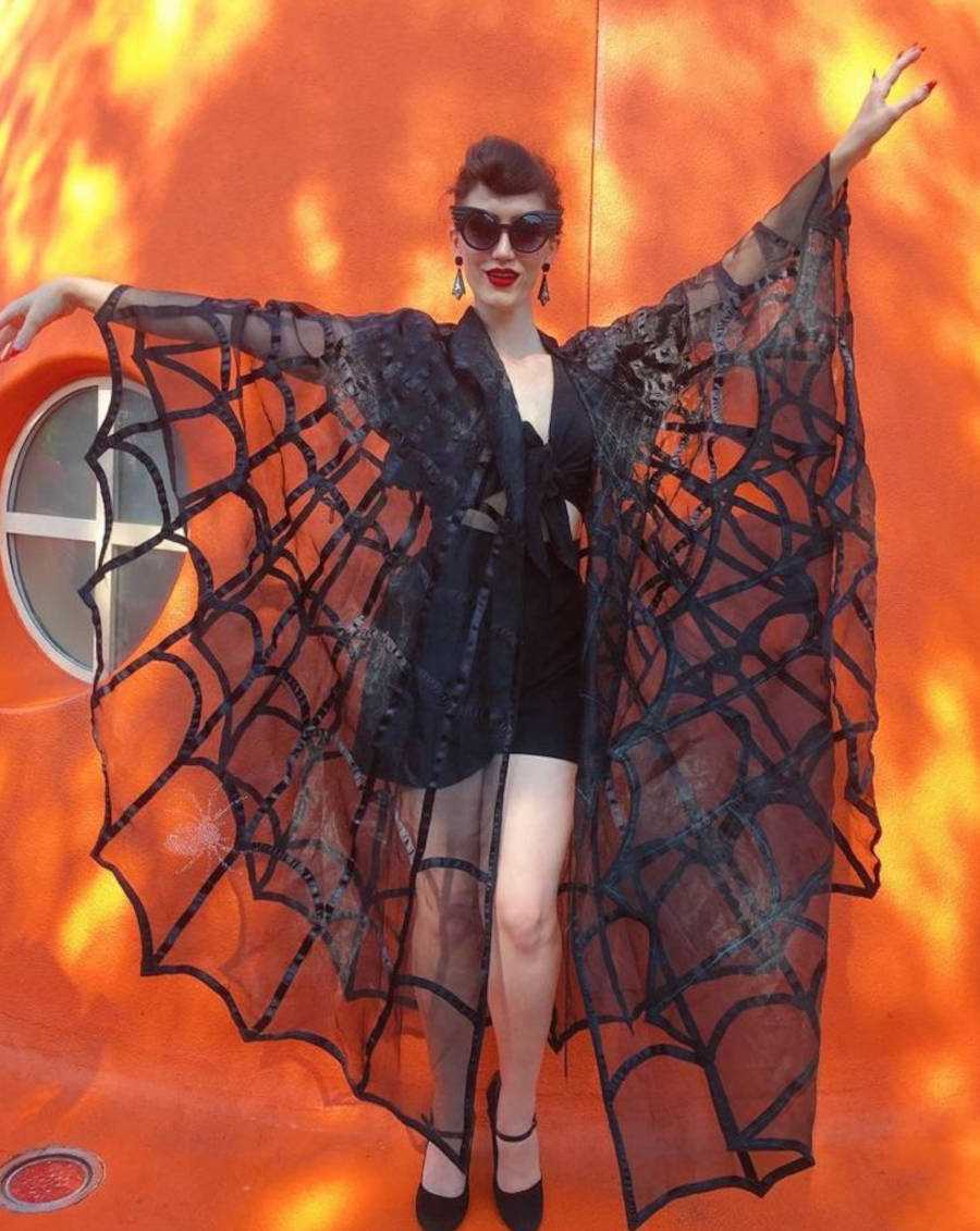 Foto de uma mulher com uma capa de teia de aranha longa, com os braços para cima e óculos escuro