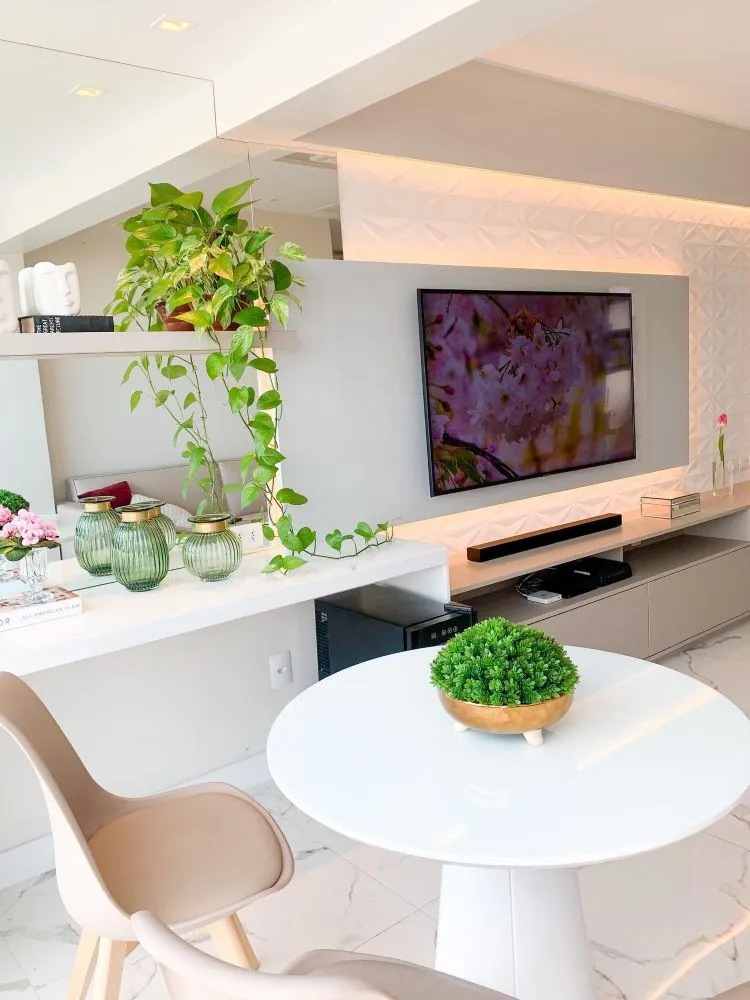 Sala clean em tons de branco com mesa redonda branca, cadeira rosa clara, televisão e jiboia pendurada em prateleira.
