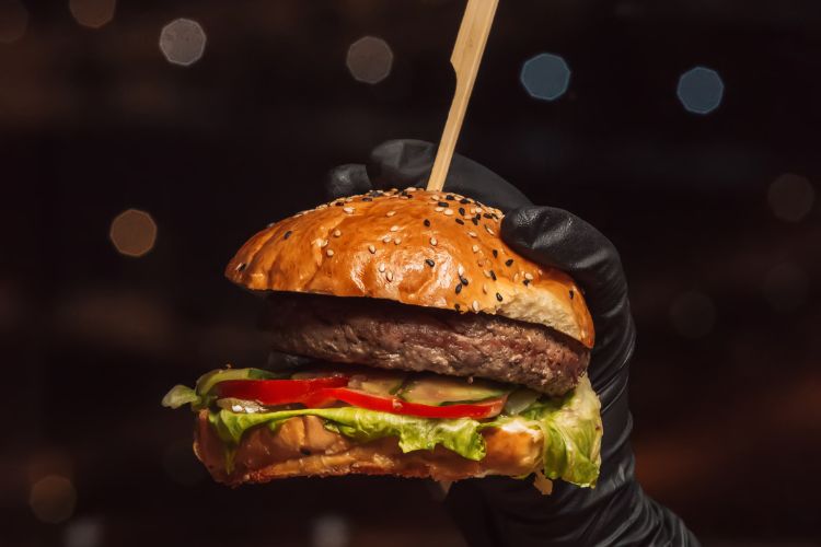 Hambúrguer clássico em pão com gergelim, recheado com alface, tomate, carne suculenta e queijo. Segurado por uma mão enluvada de preto, com fundo desfocado de luzes e um palito atravessando o lanche para sustentação.





