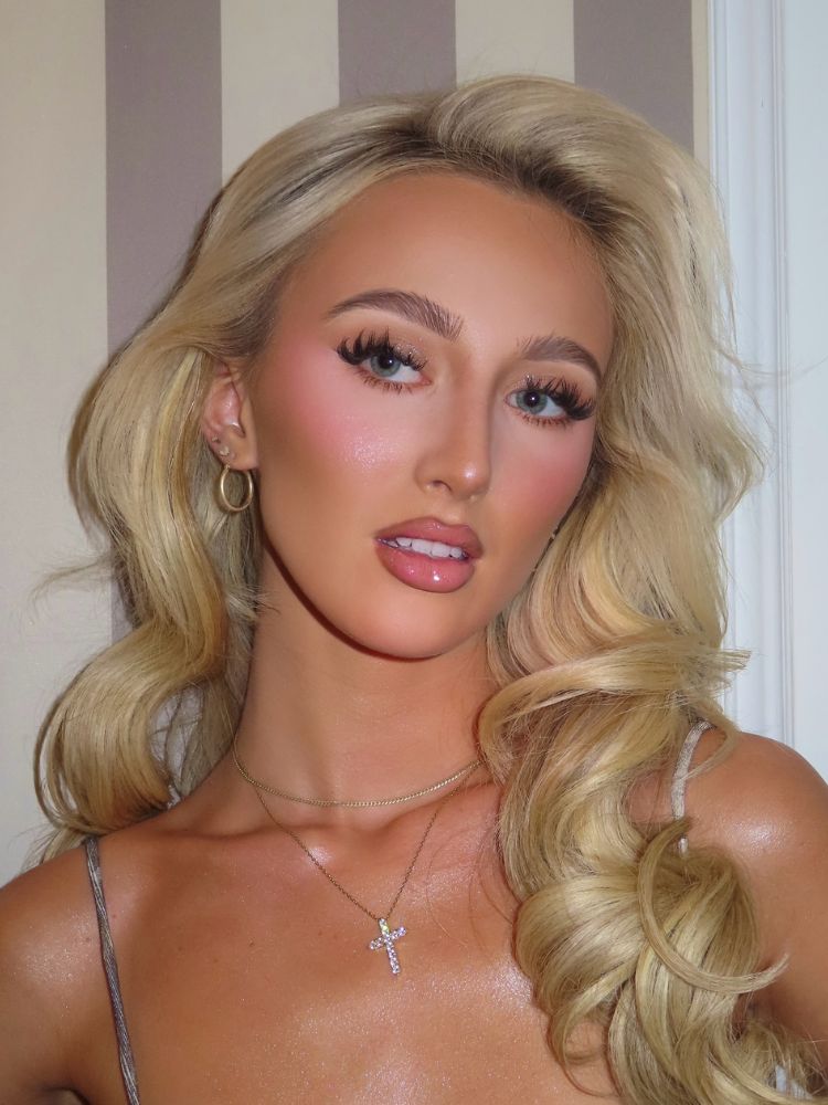 Mulher loira de pele clara com cabelo bombshell, maquiagem iluminada, blush cintilante e gloss 