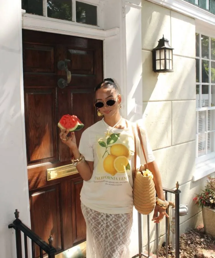 Mulher sorrindo em uma porta, usando uma camiseta "California Lemon" e uma saia de renda transparente. Ela segura uma fatia de melancia e uma bolsa de palha, usando óculos escuros e joias douradas.